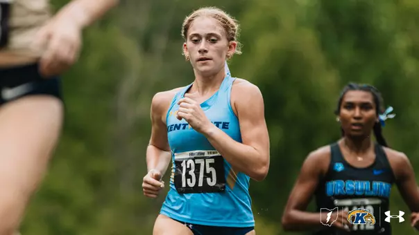 A Kent State cross country runner wearing bib No. 1375 competes in a race, running past a blurred competitor in the foreground. An Ursuline runner is visible behind her on the course.