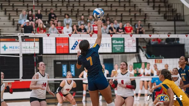 Kent State volleyball player No. 7 leaps at the net to spike the ball during a match against Youngstown State, with opposing blockers No. 14 and No. 6 in white jerseys positioned to defend.