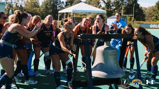 Kent State field hockey players in navy jerseys gather around and ring the "Starner Victory Bell" on the sideline in celebration after a win, smiling and cheering together on the turf field.
