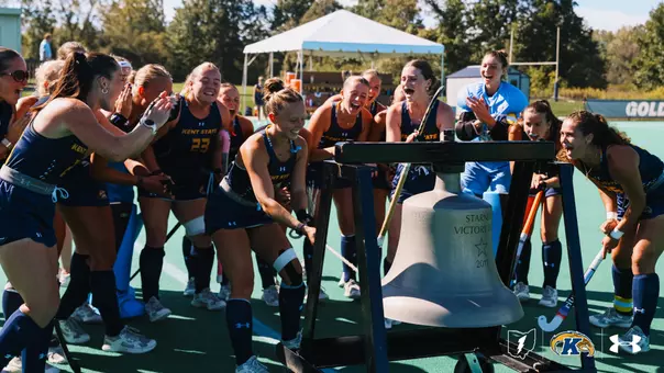 Kent State field hockey players in navy jerseys gather around and ring the "Starner Victory Bell" on the sideline in celebration after a win, smiling and cheering together on the turf field.