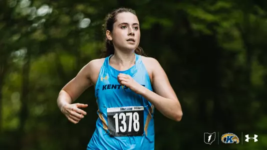 A Kent State cross country runner wearing bib number 1378 races through a wooded course in a light blue Kent State singlet, with dense green trees blurred in the background