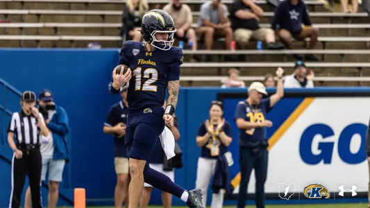 A Kent State football player wearing jersey number 12 runs down the field holding a football while pumping his left fist in celebration.