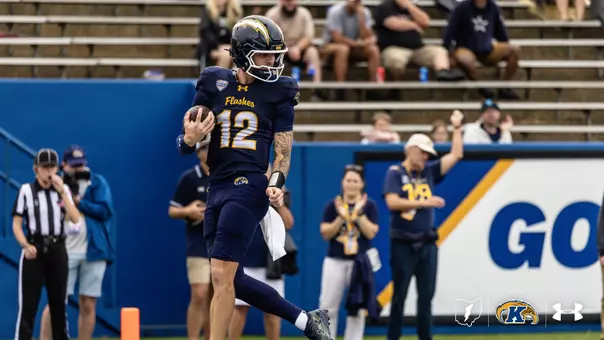 A Kent State football player wearing jersey number 12 runs down the field holding a football while pumping his left fist in celebration.