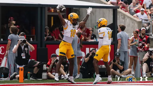 A Kent State football player wearing jersey number 18 leaps into the air with both arms raised holding a football, celebrating an interception in the end zone alongside a teammate wearing number 14.