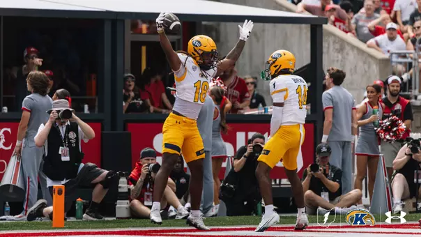 A Kent State football player wearing jersey number 18 leaps into the air with both arms raised holding a football, celebrating an interception in the end zone alongside a teammate wearing number 14.