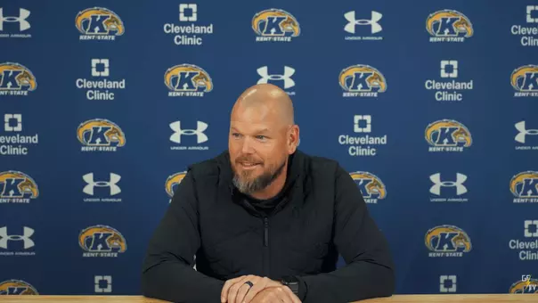 A smiling man with a beard sits at a wooden table during a press conference in front of a blue Kent State backdrop.