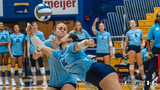 Two Kent State volleyball players collide while attempting to play a low ball during a match, with teammates watching from the sideline behind them."