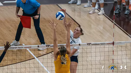 Kent State player in a white jersey sets the ball at the net during a road match at Virginia Tech, with an opposing blocker reaching up and a line judge signaling with a red flag in the background."