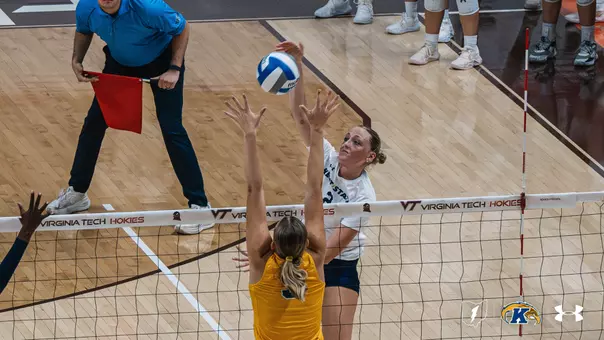 Kent State player in a white jersey sets the ball at the net during a road match at Virginia Tech, with an opposing blocker reaching up and a line judge signaling with a red flag in the background."