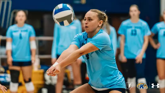 Kent State libero digs a volleyball, arms extended low as the ball approaches, with teammates watching in the background.