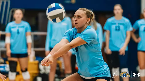 Kent State libero digs a volleyball, arms extended low as the ball approaches, with teammates watching in the background.