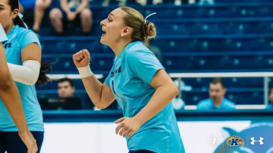Kent State player pumps her fist and shouts in celebration during a match, with teammates and fans in the stands behind her.