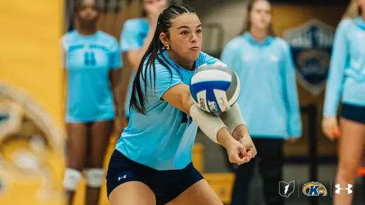 Kent State player number 12 digs the ball with a forearm pass, teammates watching in the background.