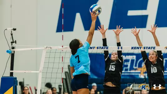 Kent State player No. 7 tips the ball over the net as two Colorado State blockers, wearing black, reach up to defend at the net.