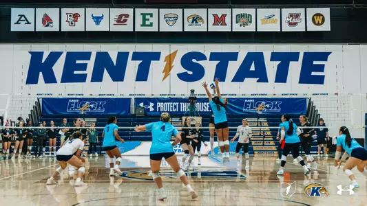 Wide shot of a Kent State volleyball match in action under the MAC conference team banners, with a Kent State player (#8) jumping to set the ball.