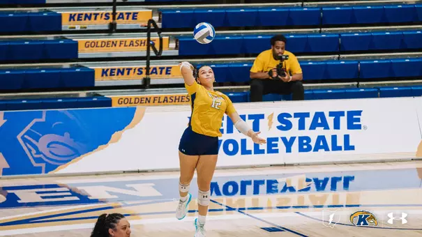 Kent State player No. 12, in gold, leaps into the air to serve, ball tossed high above her.