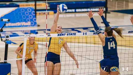 Kent State middle blocker No. 6 swings for a kill at the net as Akron's No. 14 jumps to block, with a Kent State teammate watching from behind.