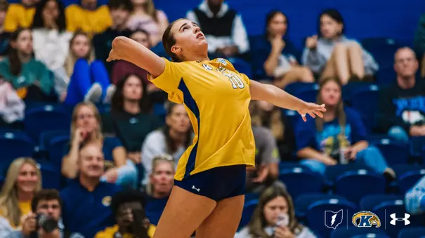 A Kent State volleyball player wearing a gold jersey with the number 10 winds up to serve, arm drawn back, eyes tracking the ball, with fans seated in the blue-and-gold stands blurred behind her.
