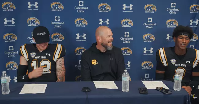 Press conference scene featuring two Kent State football players and a coach seated at a table in front of a branded media backdrop, smiling and talking