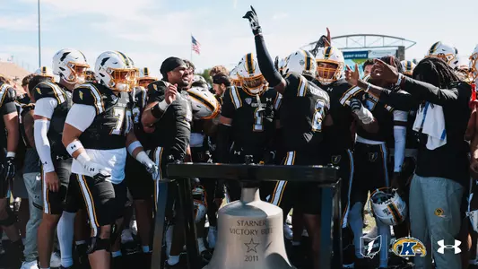 Kent State football players gathering in celebration around the Starner Victory Bell on the field.