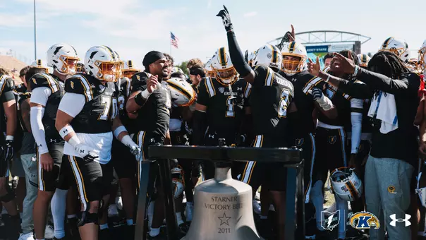 Kent State football players gathering in celebration around the Starner Victory Bell on the field.