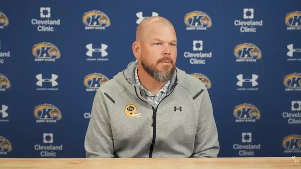A bald coach with a beard wearing a gray Kent State hooded jacket seated in front of a branded media backdrop.