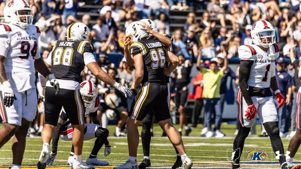 Action photo of Kent State football players embracing in celebration while opposing Ball State players stand nearby on the field.