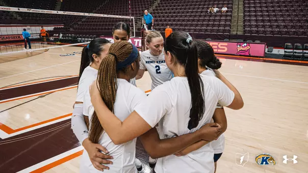 Kent State volleyball players huddle together with arms around each other's shoulders before a match at Virginia Tech's arena, with the Hokies' logo and empty maroon seating visible in the background. Player No. 2 stands in the center of the huddle wearing a white Kent State jersey.