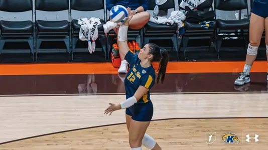 Kent State volleyball student-athlete Catie Patterson executes a jump serve, sending the ball into the air on the road at an opponent's gym, with a teammate's legs visible in the background.