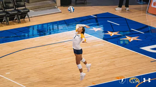 Kent State volleyball player Mercedez Brown leaps into the air to execute a jump serve, tracking the ball above her on a star-patterned away court.