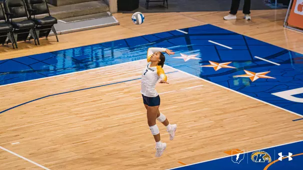 Kent State volleyball player Mercedez Brown leaps into the air to execute a jump serve, tracking the ball above her on a star-patterned away court.
