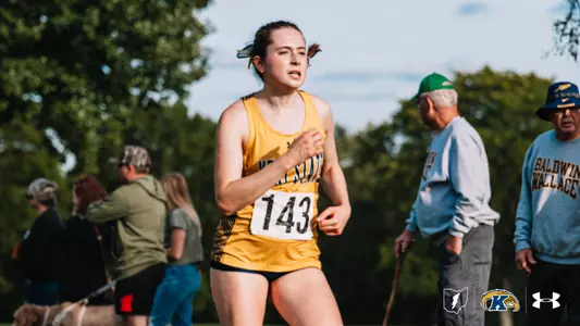 Kent State cross country runner wearing bib No. 143 competes during a race, with spectators, a Baldwin Wallace fan, and dogs visible along the course in the background.