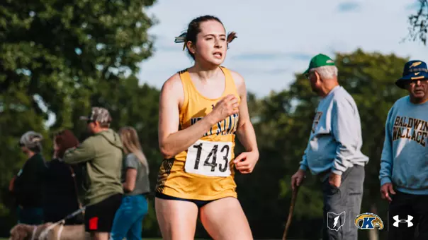Kent State cross country runner wearing bib No. 143 competes during a race, with spectators, a Baldwin Wallace fan, and dogs visible along the course in the background.