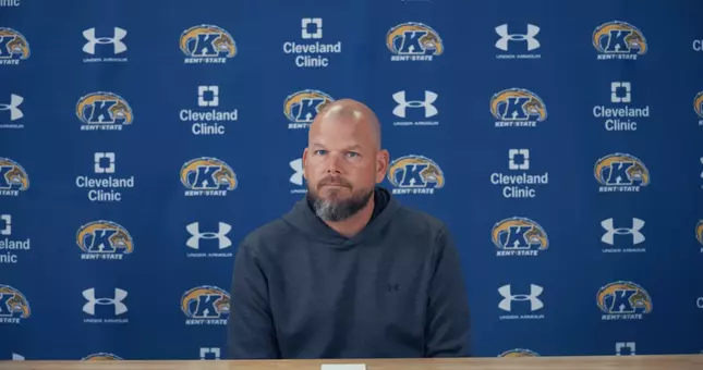 A coach sitting at a press conference table in front of a blue Kent State step-and-repeat backdrop wearing a dark gray hoodie.