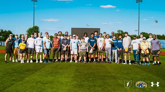 A large group photo on a baseball field of Kent State University athletes and staff standing in a row, along with a young boy in a green Kent State jersey (Team IMPACT signee Alex) standing among them. Several people hold softball gloves and bats. Behind them is a scoreboard, light towers, and trees under a blue sky with scattered clouds. Logos for the state of Ohio, Kent State Golden Flashes, and Under Armour appear in the bottom right corner.