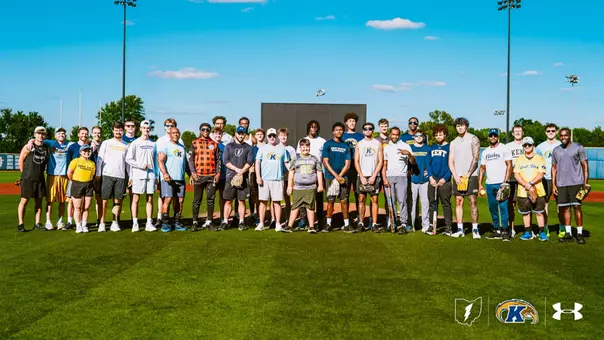A large group photo on a baseball field of Kent State University athletes and staff standing in a row, along with a young boy in a green Kent State jersey (Team IMPACT signee Alex) standing among them. Several people hold softball gloves and bats. Behind them is a scoreboard, light towers, and trees under a blue sky with scattered clouds. Logos for the state of Ohio, Kent State Golden Flashes, and Under Armour appear in the bottom right corner.