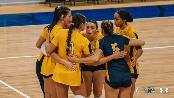 Kent State volleyball players huddle on the court with arms around each other's shoulders, wearing gold and navy jerseys.