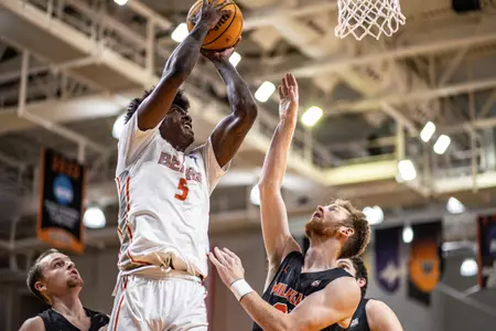 Mercer men's basketball player wearing a white jersey in the air about to dunk the basketball while a defender's hand is in the air trying to block it