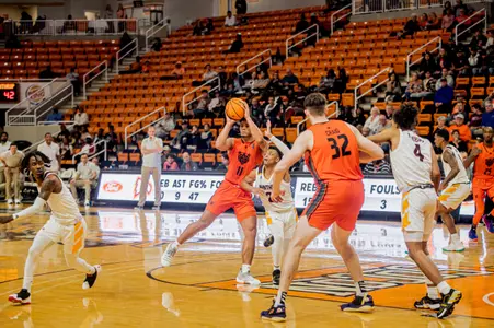 Mercer men's basketball player wearing an orange jersey holding the ball above his head looking to pass the ball while a defender in white tries to block it