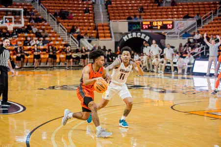 Mercer men's basketball player wearing an orange jersey dribbling the ball inside the arc with a defender in white trying to stop him