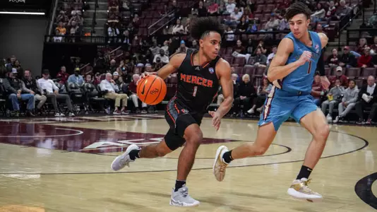 Mercer men's basketball player wearing a black jersey running down the court with the ball in his hand while a defender runs alongside him