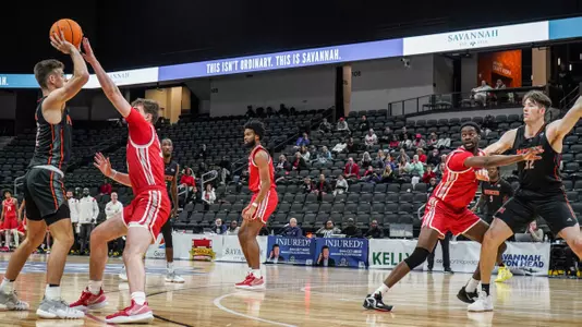 Mercer men's basketball player wearing a black jersey begins to throw the basketball over his head to a teammate while both players are being guarded by defenders wearing red.