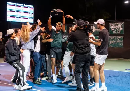 Matt Forbes raises the 2025-26 Men's Tennis Big Ten Tournament trophy while getting swarmed by teammates