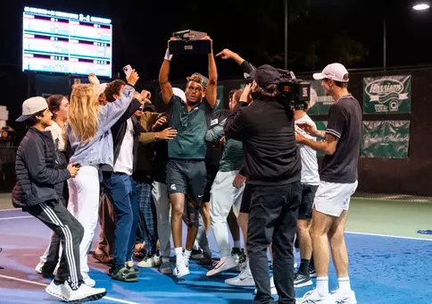 Matt Forbes raises the 2025-26 Men's Tennis Big Ten Tournament trophy while getting swarmed by teammates