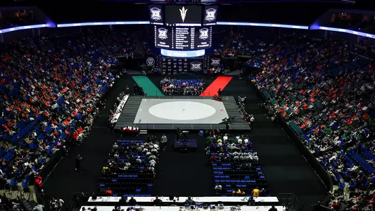 Wide overhead view of BOK Center set up for Big 12 wrestling tournament, featuring a single gray mat in the center, a large hanging scoreboard with conference logos above it, and spectators filling the surrounding seats on all sides.