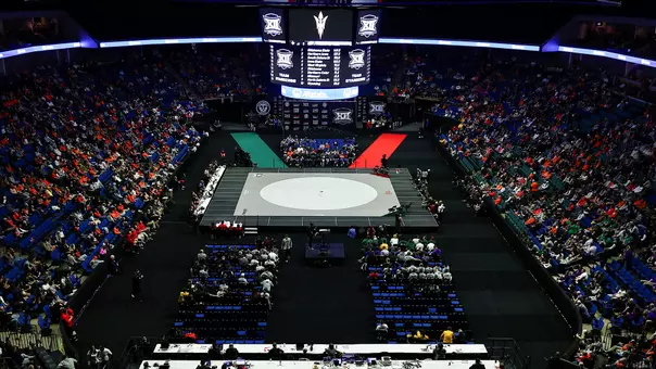 Wide overhead view of BOK Center set up for Big 12 wrestling tournament, featuring a single gray mat in the center, a large hanging scoreboard with conference logos above it, and spectators filling the surrounding seats on all sides.