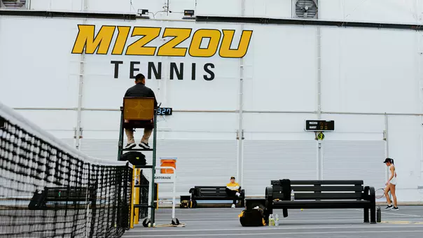 Indoor tennis court at the Mizzou Tennis facility with a large “MIZZOU TENNIS” sign on the back wall. An official sits in a high umpire chair near the net while a player on the far right prepares between points. Benches, tennis bags, water bottles, and a Gatorade cooler line the sideline, and a scoreboard in the background shows Mizzou leading 5–0.