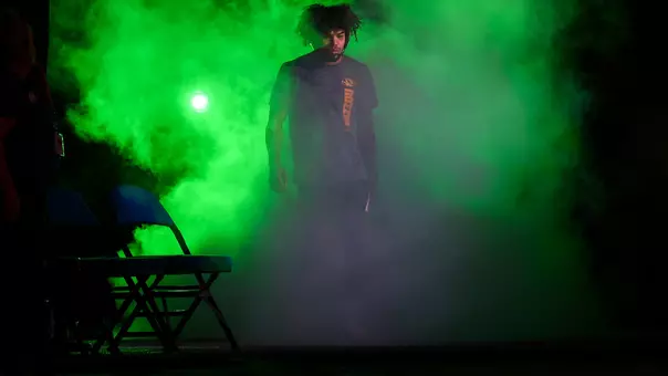 A male athlete walks through thick green smoke during a dramatic arena entrance. A bright green light glows behind him, silhouetting his figure as folding chairs sit in the foreground on a dimly lit stage.