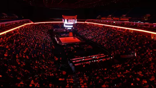 Wide overhead view of a wrestling arena inside the BOK Center during the Big 12 Championships. The arena is dimly lit with dramatic red lighting around the concourse and ceiling trusses. A single wrestling mat sits illuminated at the center of the floor beneath a large scoreboard, surrounded by a packed crowd filling the lower bowl while the upper sections fade into darkness. The scene creates a high-energy, championship atmosphere focused on the mat in the middle of the arena.