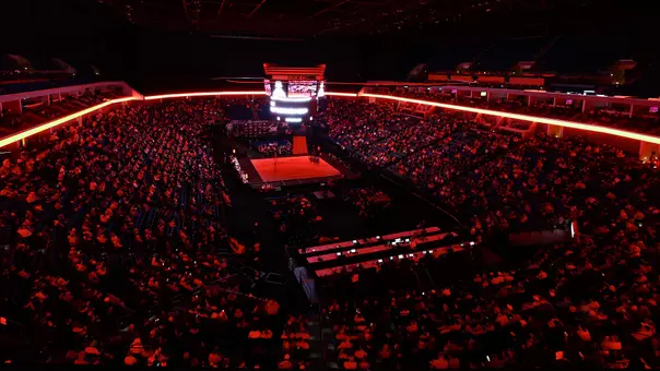 Wide overhead view of a wrestling arena inside the BOK Center during the Big 12 Championships. The arena is dimly lit with dramatic red lighting around the concourse and ceiling trusses. A single wrestling mat sits illuminated at the center of the floor beneath a large scoreboard, surrounded by a packed crowd filling the lower bowl while the upper sections fade into darkness. The scene creates a high-energy, championship atmosphere focused on the mat in the middle of the arena.
